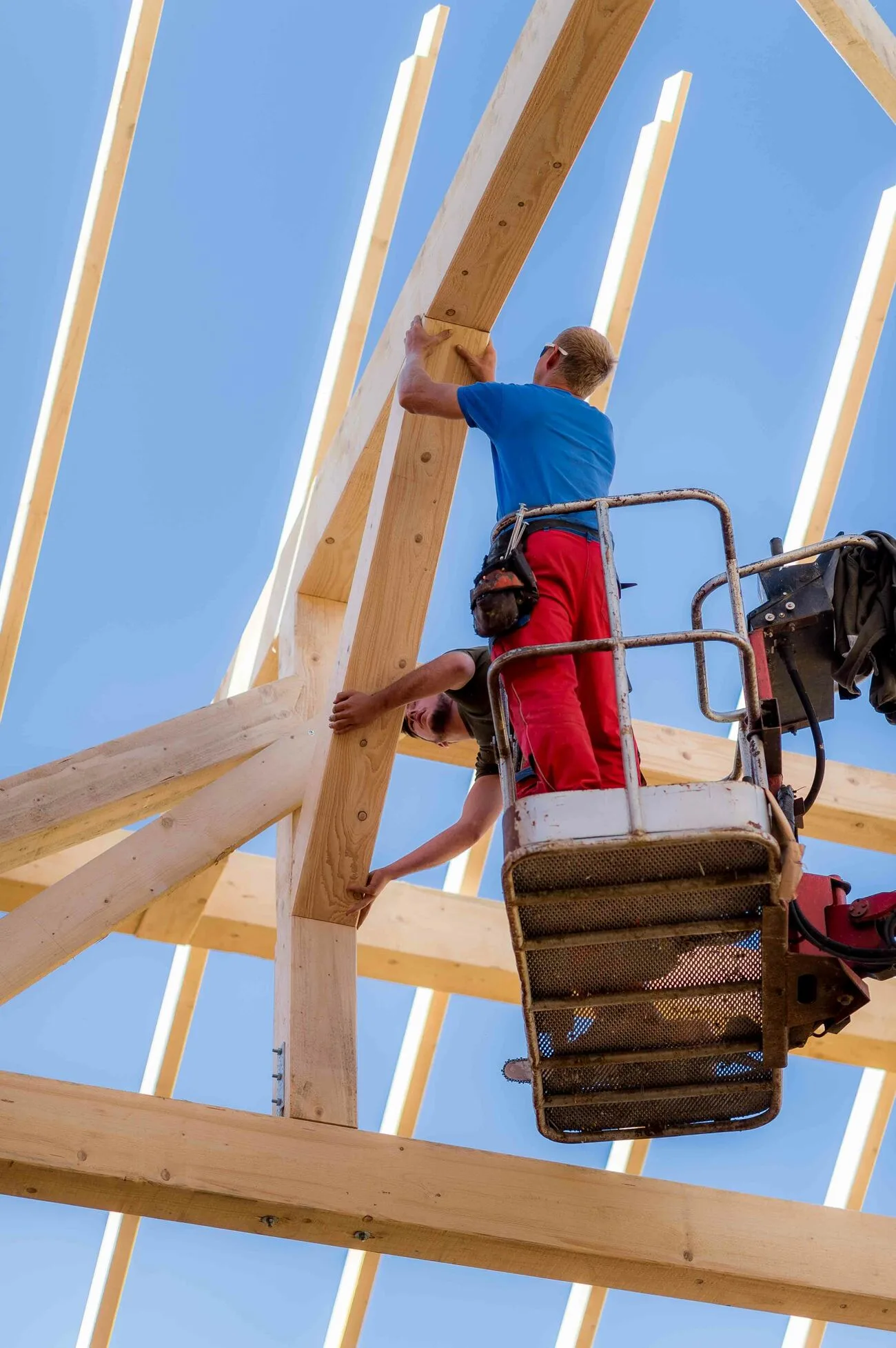 Zimmerer in Firmenkleidung im Arbeitskorb einer Hebebühne zwischen den Sparren eines Dachstuhls