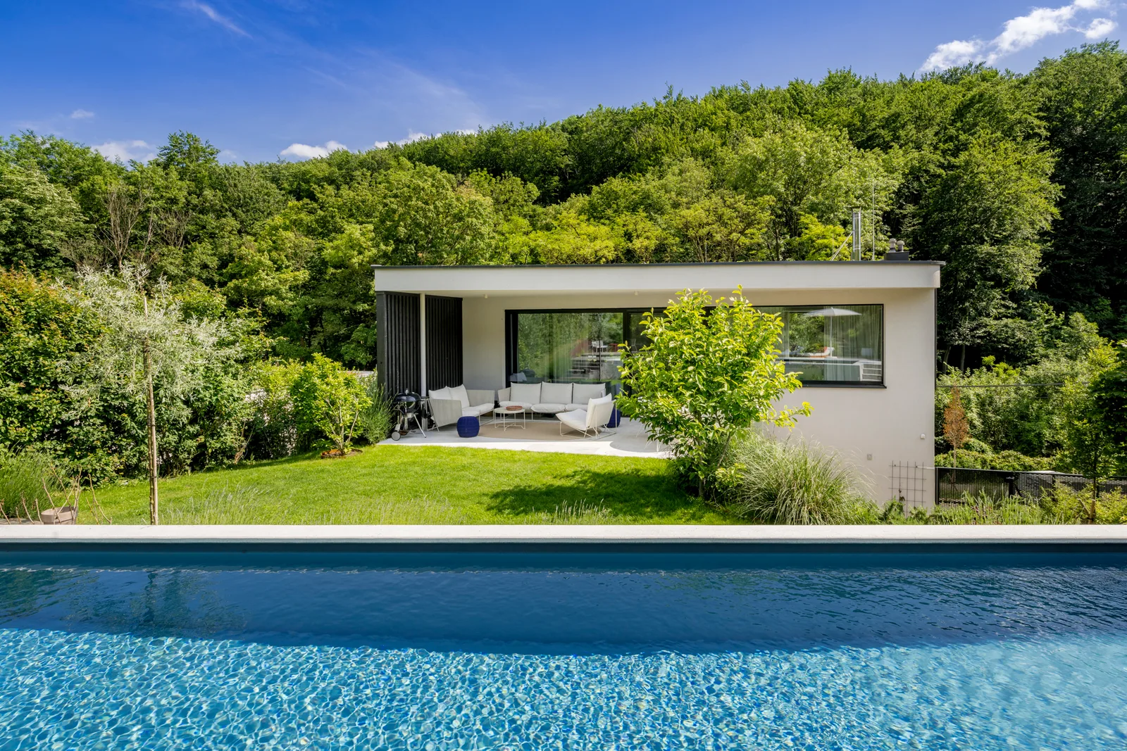 Gartenpavillon mit Flachdach und großen Fenstern hinter einem Pool