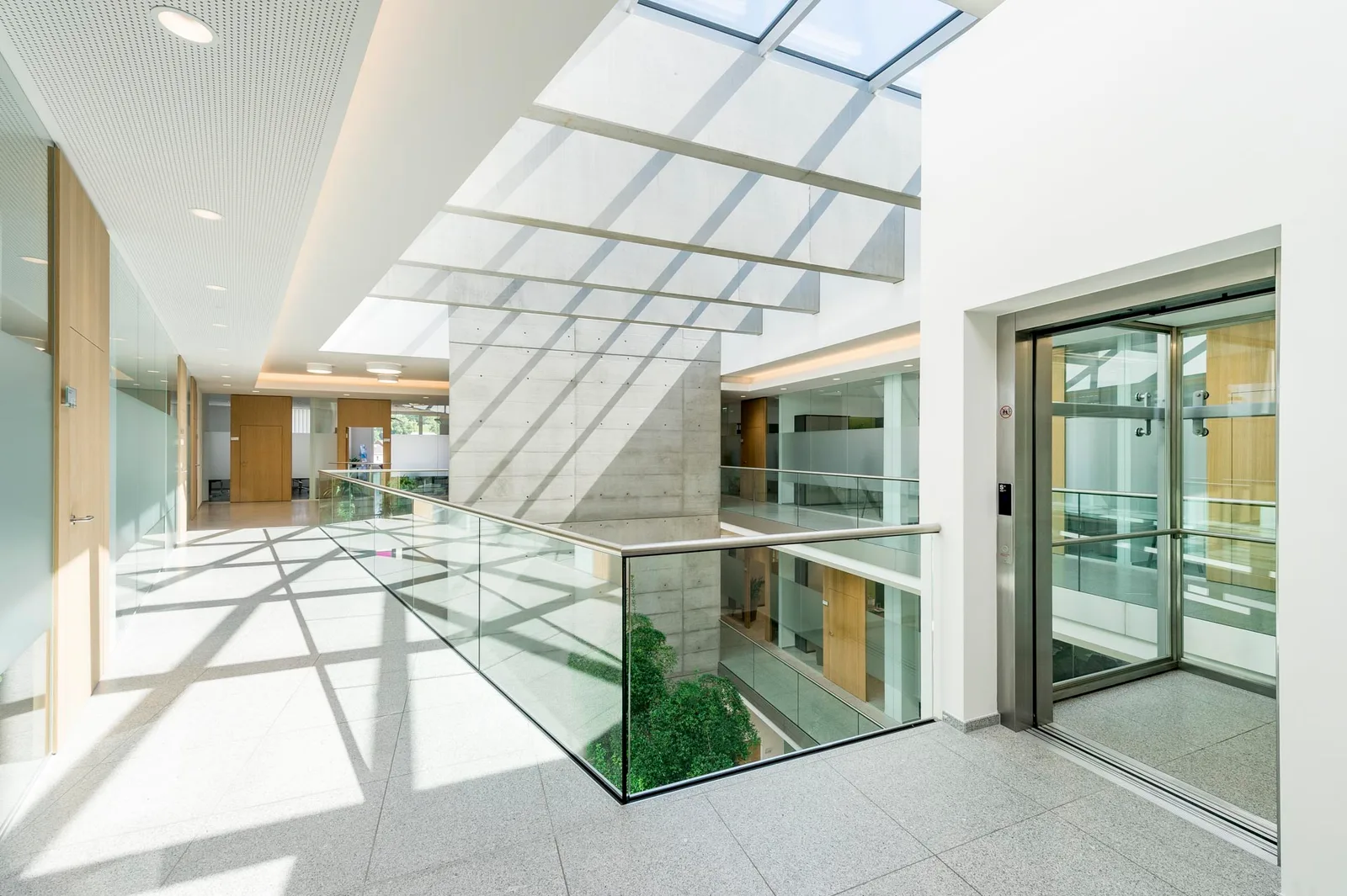 Heller Gang im Bürogebäude unter einem Glasdach, daneben ein gläserner Lift